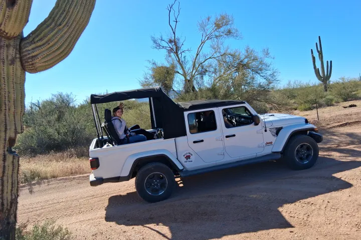 White off-road vehicle driving on desert trail near cacti under clear blue sky.
