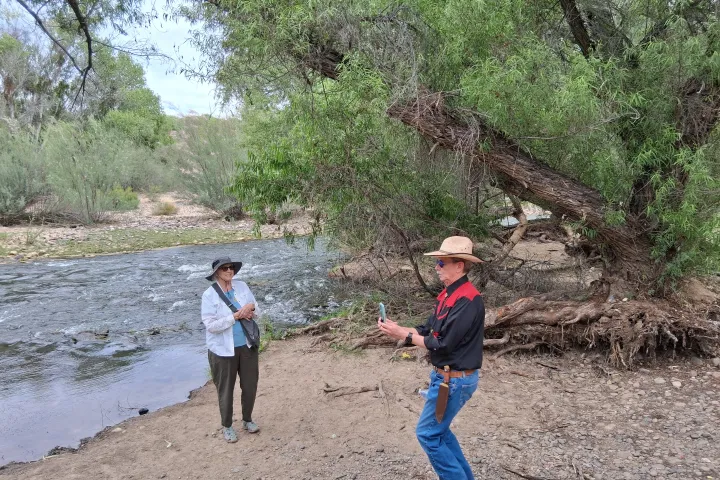 Two people enjoying nature near a river with trees and rocky shore.