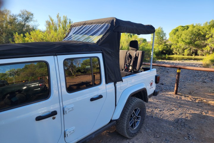 White Jeep with a black soft top, parked on a dirt road near trees.