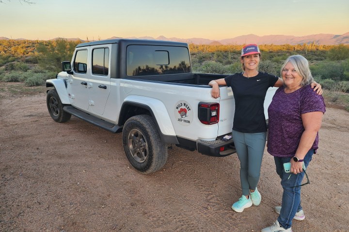 Two women smiling beside a white jeep in a desert landscape at sunset.