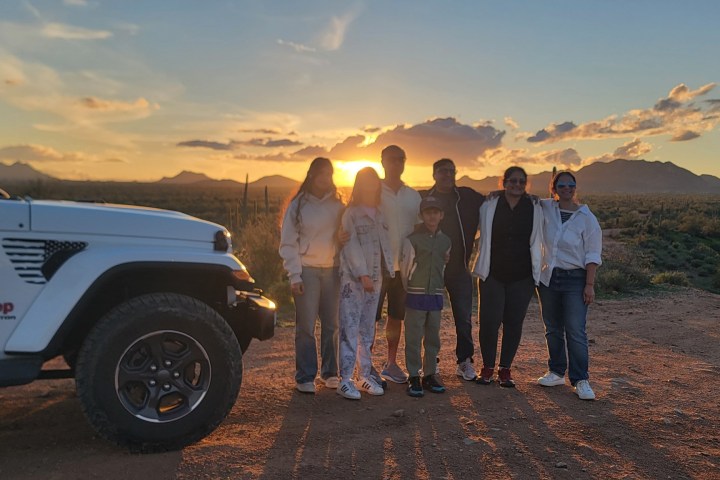 Group of people standing near a white Jeep with a sunset backdrop.