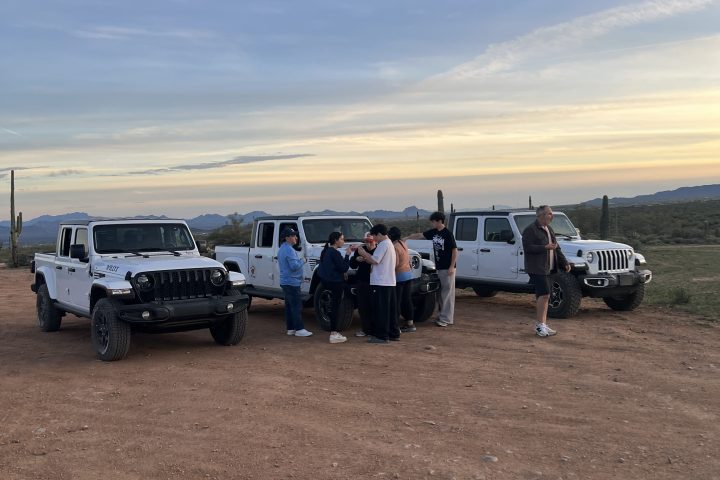 Group of people standing between three parked white SUVs on a dirt road, with a scenic horizon in the background.
