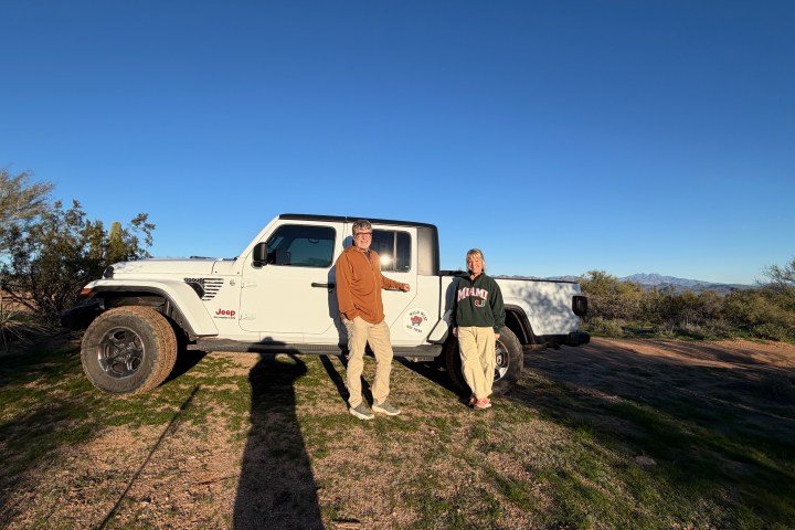 Two people standing beside a white Jeep in a desert landscape under a clear blue sky.
