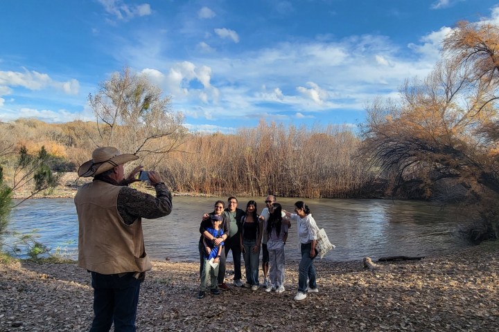 Person taking a photo of a group by a river surrounded by trees under a blue sky with clouds.