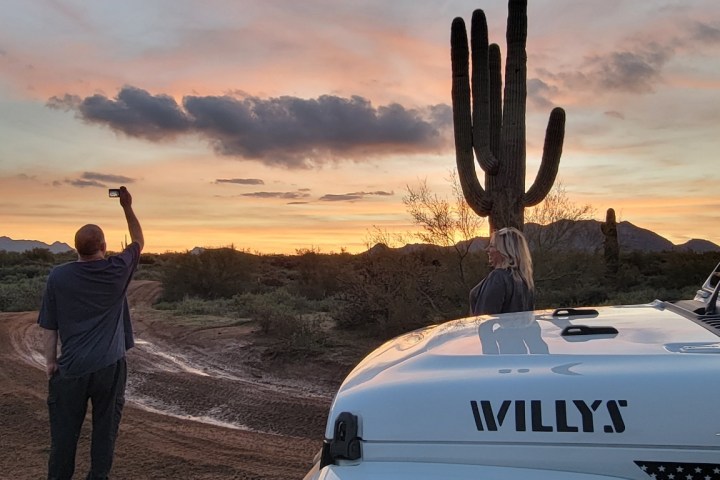 Person photographing sunset by a cactus, with a Jeep in the foreground.