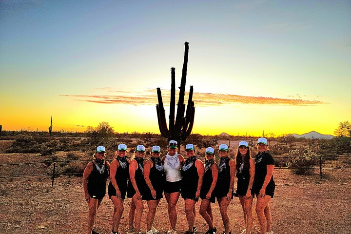 Group of women in matching outfits posing in front of a desert sunset with a large cactus.