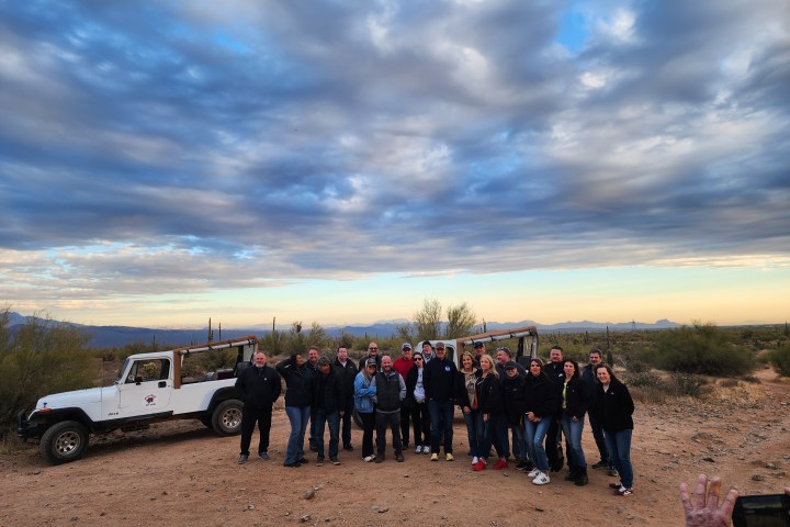 Group of people posing in a desert with a white Jeep and dramatic cloudy sky.