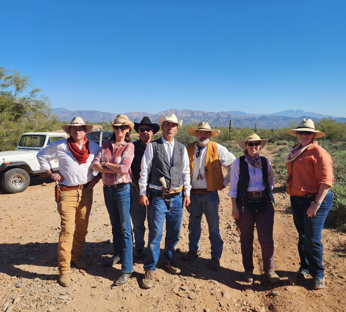 Group of people in cowboy attire standing on a rocky desert path with blue sky.